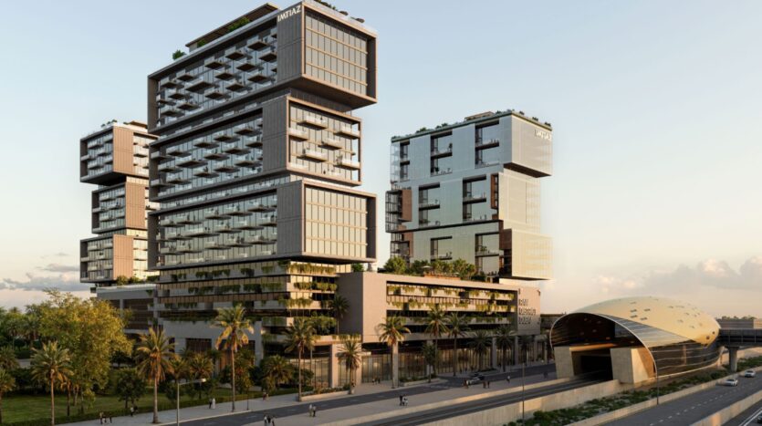 Des gratte-ciel modernes aux façades de verre empilées et en surplomb se dressent à côté d'une station de métro futuriste de forme ovale près de Raw District by Imtiaz ; des palmiers bordent la rue en face, avec quelques personnes marchant à proximité.