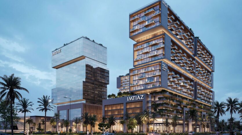 Raw District by Imtiaz est un complexe immobilier moderne composé de deux tours principales avec des façades en verre et de grandes terrasses, entourées de palmiers. Le nom "IMTIAZ" est affiché sur les deux tours. La scène du crépuscule est éclairée par des lumières intérieures accueillantes.