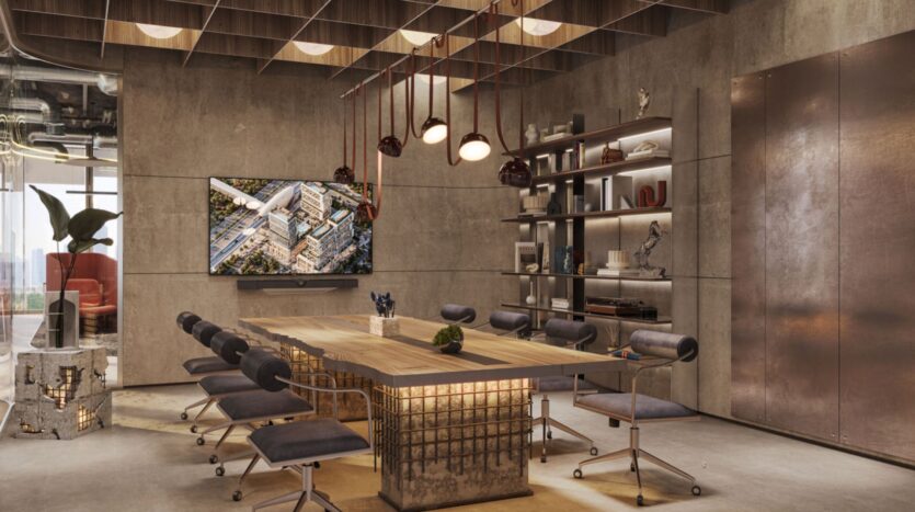 Salle de conférence moderne avec des murs en béton, une grande table en bois Raw District by Imtiaz, huit chaises rembourrées sur roulettes, des lampes suspendues, des étagères décorées, une télévision murale et une grande fenêtre avec vue sur la ville.
