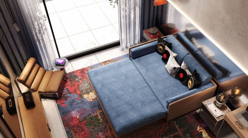 Le salon confortable du Raw District by Imtiaz est composé d'un canapé-lit bleu, d'un tapis à motifs colorés, de coussins en forme de disques vinyles, d'une chaise longue beige et de portes coulissantes donnant sur un balcon. Divers objets de décoration et gadgets ajoutent à l'atmosphère vibrante.