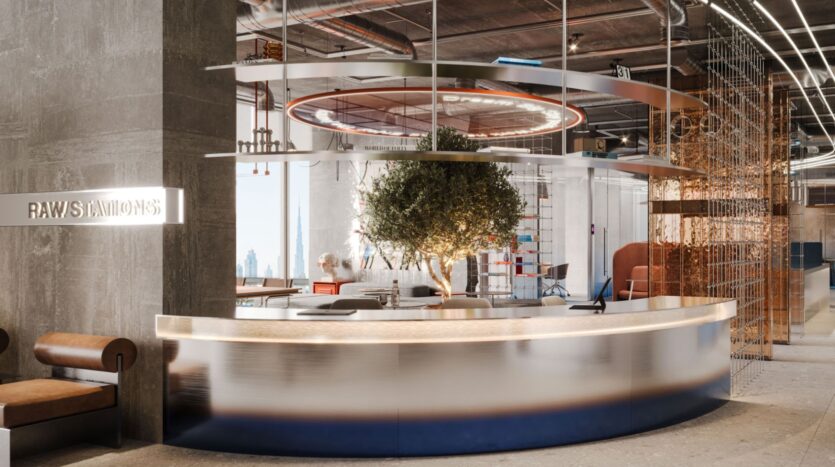 Une réception de bureau moderne avec un bureau incurvé, un arbre au centre, un éclairage élégant et un décor industriel avec des tuyaux apparents et des murs en béton. Un panneau indique "RAW STATION by Raw District by Imtiaz.
