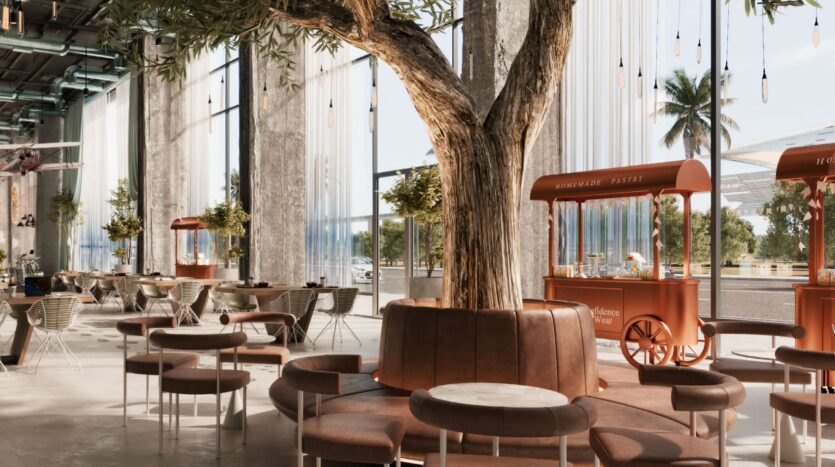 Le spacieux café moderne Raw District by Imtiaz est doté de grandes fenêtres, d'une lumière naturelle abondante et d'un grand arbre au centre duquel se trouvent des sièges bruns. Les chariots de nourriture orange et la verdure extérieure brillent à travers les vitres, tandis que les tables et les chaises sont judicieusement disposées.