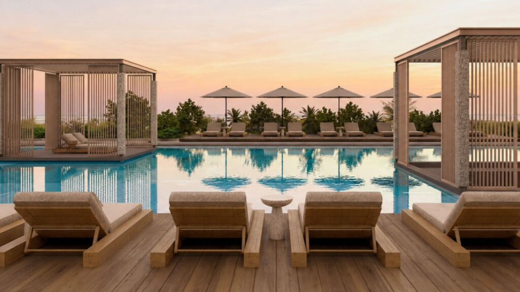 Scène luxueuse au bord de la piscine au coucher du soleil au Raw District by Imtiaz, avec des chaises longues en bois, des cabanas ombragées et des parasols se reflétant dans une piscine calme, entourée de verdure et d'un ciel rose orangé en arrière-plan.