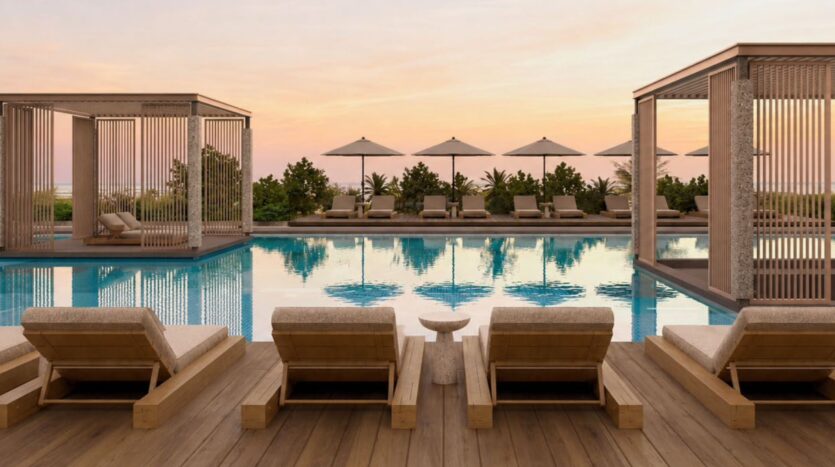 Scène luxueuse au bord de la piscine au coucher du soleil au Raw District by Imtiaz, avec des chaises longues en bois, des cabanas ombragées et des parasols se reflétant dans une piscine calme, entourée de verdure et d'un ciel rose orangé en arrière-plan.