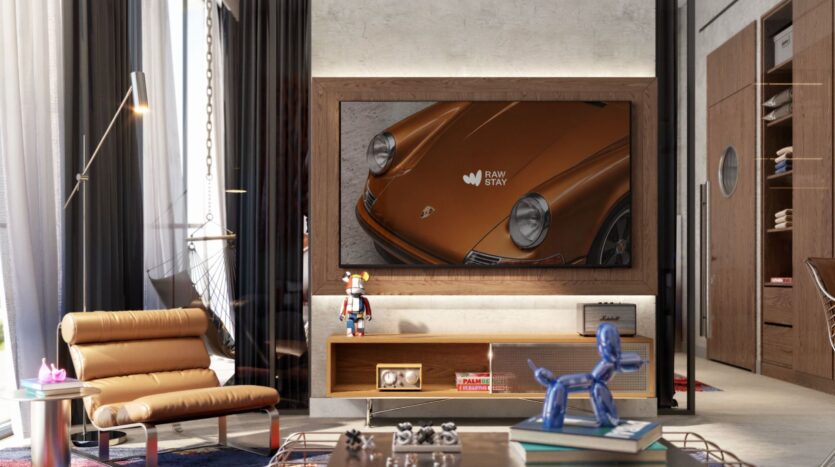 Un salon moderne avec un fauteuil en cuir fauve, un meuble TV en bois, des figurines décoratives et un grand tableau mural représentant l'avant d'une voiture de collection orange sur lequel est écrit "RAW STAY", clin d'œil à Raw District d'Imtiaz.
