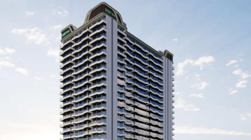 Un grand immeuble moderne, Reef 996, avec de nombreux balcons et baies vitrées, se dresse sur un ciel bleu parsemé de nuages. La structure présente des angles incurvés et des panneaux de signalisation sur le toit.