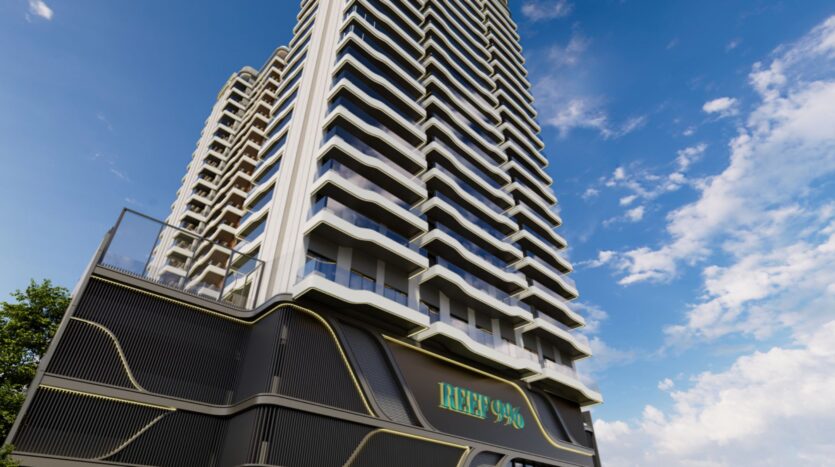 Immeuble moderne de grande hauteur avec des balcons ondulés, vu d'en bas sur un ciel bleu avec des nuages. Un panneau indiquant "REEF 996" se trouve à l'entrée de l'immeuble.