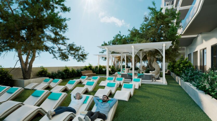 Des personnes se détendent sur des chaises longues aux coussins turquoise près de l'espace extérieur herbeux du Reef 996, entouré d'arbres et de verdure. Une pergola blanche avec des chaises suspendues se trouve à proximité, adossée à un bâtiment moderne sous un ciel ensoleillé.