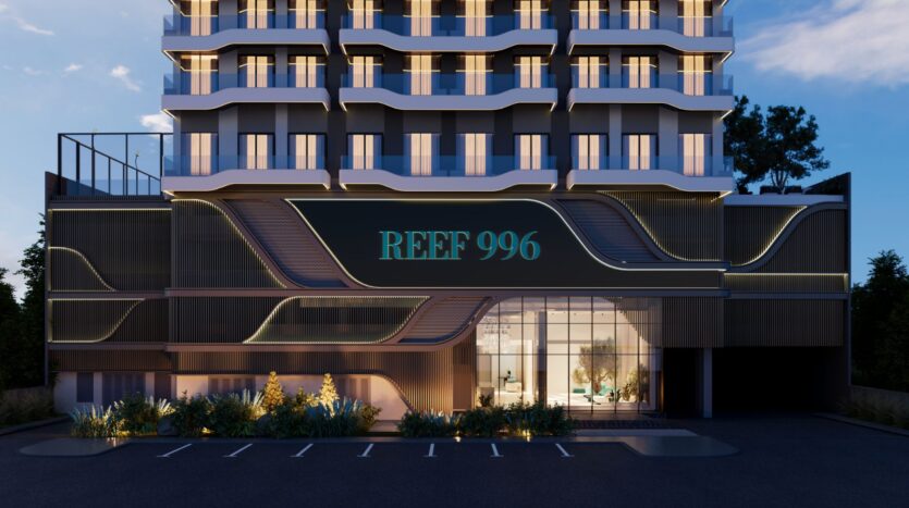 Façade d'un bâtiment moderne au crépuscule avec des fenêtres éclairées ; une grande enseigne "Reef 996" au-dessus de l'entrée en verre. Les lignes architecturales courbes et l'aménagement paysager se distinguent, avec des places de parking vides devant Reef 996.