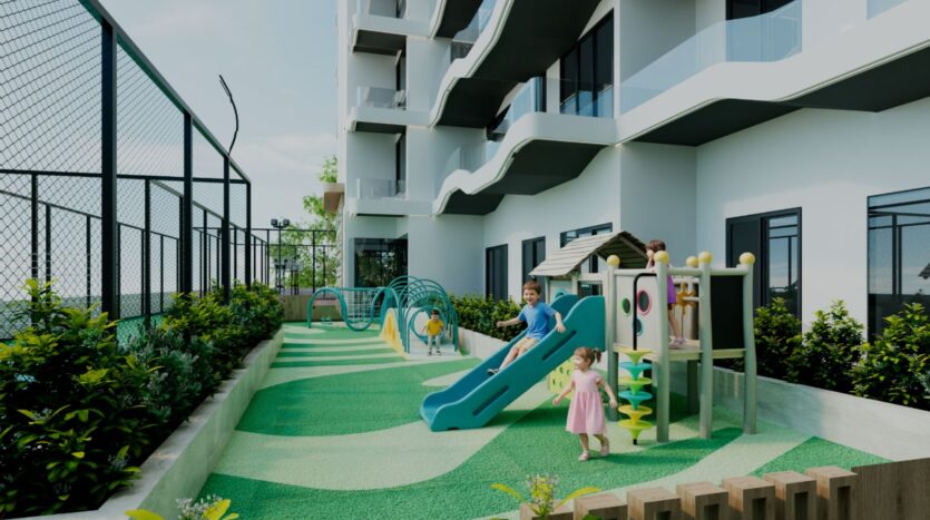 Des enfants jouent sur une petite aire de jeux avec un toboggan bleu, des structures d'escalade et des tunnels dans un espace extérieur à côté de l'immeuble d'appartements moderne Reef 996 avec des balcons et des garde-corps en verre. L'aire de jeu est bordée de verdure.