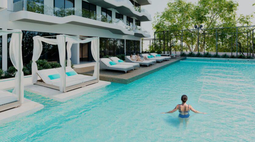 Une femme se tient dans une piscine d'un bleu limpide, à proximité des 996 lits de luxe de Reef, avec des baldaquins blancs et des oreillers turquoise, à l'extérieur d'un bâtiment moderne doté de balcons et entouré d'arbres.