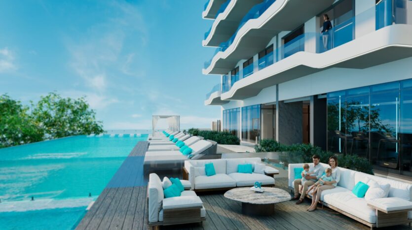 Le Reef 996 dispose d'un espace piscine moderne et luxueux, composé de chaises longues et de canapés blancs avec des coussins bleus, à côté d'un bâtiment avec des balcons en verre. Deux adultes et un enfant sont assis sur un canapé, tandis que quelqu'un se tient sur le balcon au-dessus.