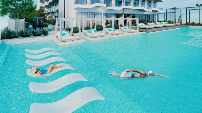 Une personne fait des longueurs dans la piscine extérieure moderne Reef 996 tandis qu'une autre se détend sur une chaise longue partiellement immergée. Des cabanons au bord de la piscine et un bâtiment contemporain complètent cette scène élégante.
