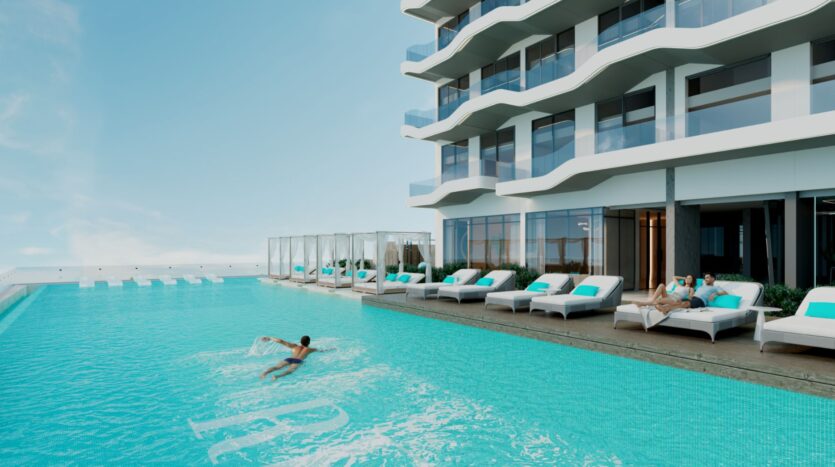 Un hôtel moderne avec balcons donne sur une grande piscine extérieure, où une personne nage et les autres se détendent sur des chaises longues blanches au bord de la piscine. Les cabanas Reef 996 aux couleurs vives et le ciel bleu forment une toile de fond parfaite.