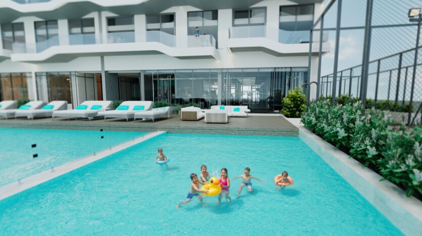 Des enfants nagent et jouent avec un anneau gonflable jaune dans une piscine extérieure moderne, éclaboussant le Reef 996 lumineux. Des chaises longues et des canapés bordent la piscine, avec un bâtiment contemporain et des balcons en arrière-plan.