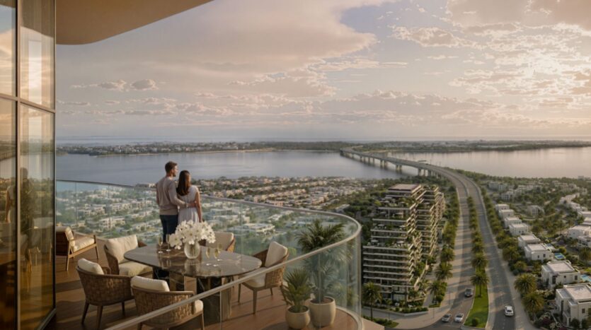 Un couple se tient sur le balcon d'une tour moderne de Rotana Residences qui surplombe une rivière, un long pont et le paysage urbain au coucher du soleil. Le balcon comprend une table à manger, des chaises, des plantes en pot et une vue étendue sur l'eau et les bâtiments.