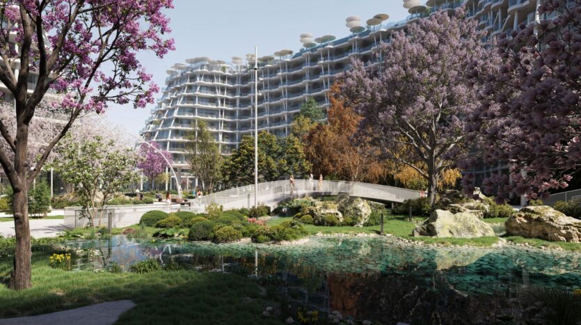 Les jardins de Sakura sont composés d'arbres en fleurs, d'un petit étang avec des rochers et d'une passerelle incurvée. À l'arrière-plan, un bâtiment moderne de plusieurs étages avec des balcons et des structures arrondies est visible sous un ciel clair.
