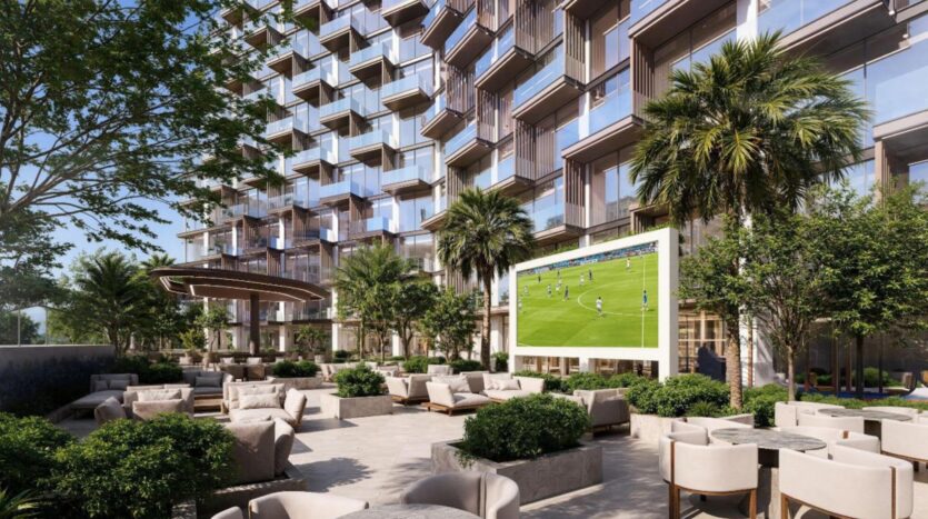 Salon extérieur moderne avec des sièges rembourrés, des plantes vertes luxuriantes et des palmiers à Winslow, situé devant un grand bâtiment avec des balcons. Un grand écran diffuse un match de football, créant ainsi une atmosphère détendue et sociale.