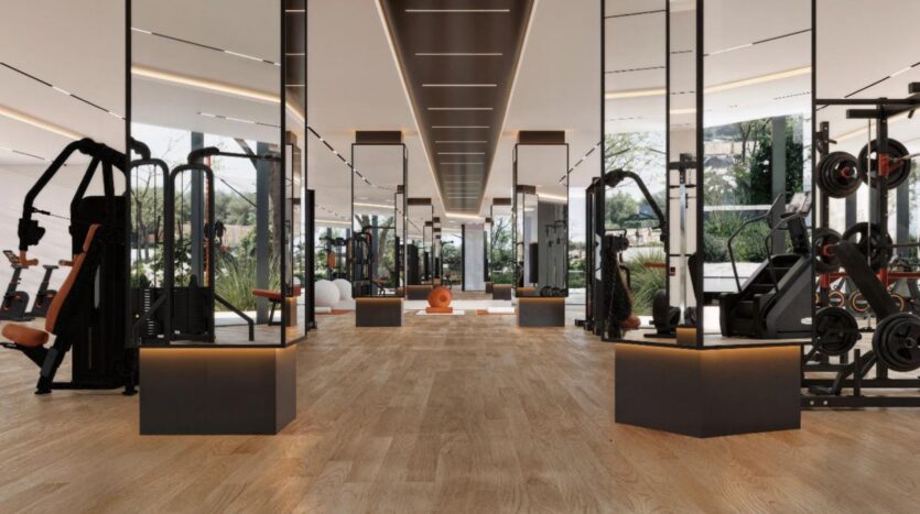 Salle de sport spacieuse et moderne de Winslow avec des sols en bois, des colonnes en miroir et de grandes fenêtres. Diverses machines de fitness et des poids sont alignés sur les côtés, tandis que la verdure est visible à l'extérieur à travers les murs en verre.