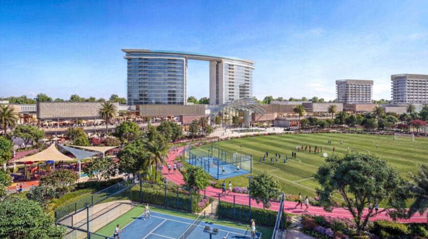 Le complexe sportif extérieur de The Woods by Sobha comprend des courts de tennis, un terrain de football, une piste de course rouge et des places assises ombragées. Des bâtiments modernes et des palmiers entourent la zone sous un ciel bleu clair.