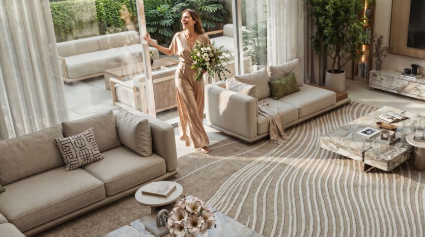Une femme tenant un bouquet de fleurs se tient près des portes vitrées ouvertes dans un salon moderne et lumineux de Tilal by Binghatti, avec des meubles aux tons neutres, un tapis à motifs et de la verdure à l'intérieur et à l'extérieur.