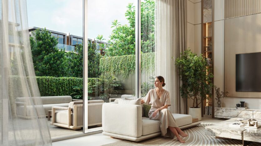 Une femme vêtue d'une robe légère est assise sur un canapé blanc moderne dans un salon lumineux de Tilal by Binghatti. De grandes portes vitrées, des plantes vertes luxuriantes à l'extérieur et la lumière naturelle remplissent l'espace contemporain et minimal.