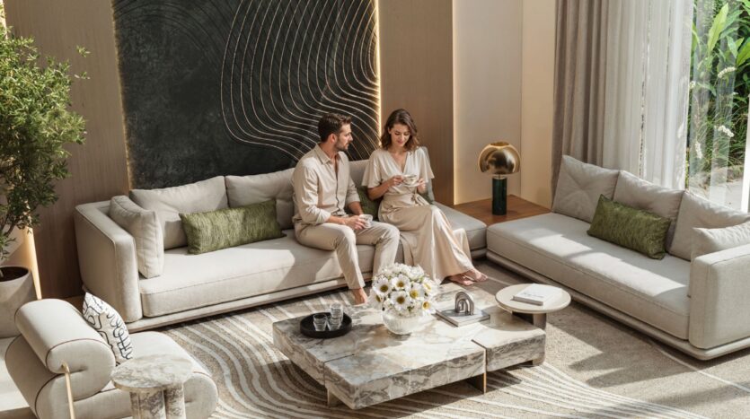 Un homme et une femme sont assis ensemble sur un canapé beige dans le salon moderne et ensoleillé de Tilal by Binghatti, qui présente des tons neutres, une table basse en marbre, des touches de vert, des œuvres d'art murales et de grandes fenêtres aux rideaux diaphanes.
