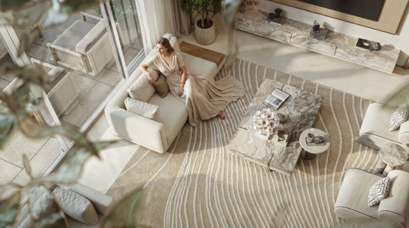 Une femme vêtue d'une longue robe beige est assise sur un canapé crème dans un salon moderne Tilal by Binghatti, doté de grandes fenêtres, de tables en marbre et d'un tapis à motifs, vu d'en haut à travers des branches feuillues.