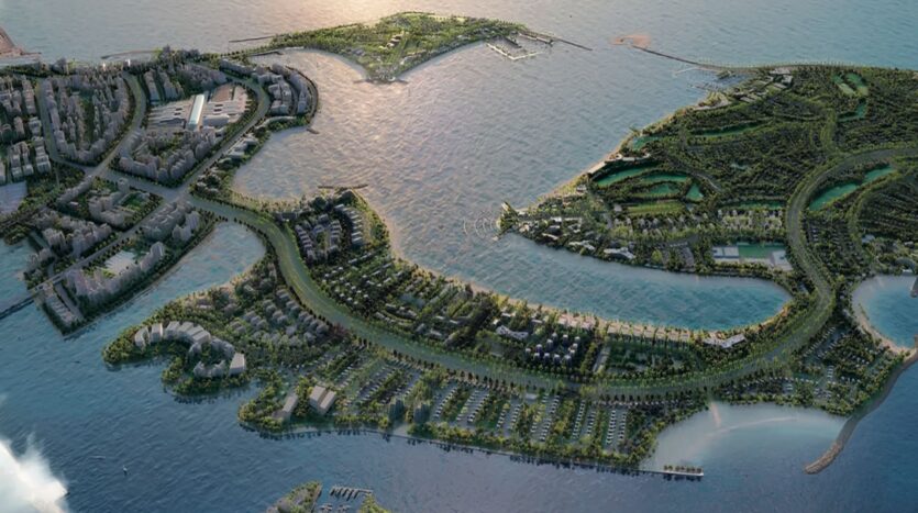 Vue aérienne d'une ville côtière avec des espaces verts luxuriants, des routes sinueuses et des bâtiments au bord de l'eau, comprenant des résidences Tivanno by AYS sur des îles reliées par des ponts, le tout entouré d'eau bleue sous un ciel nuageux.