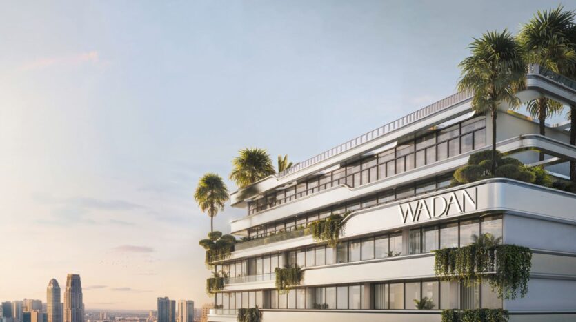Un bâtiment blanc moderne avec de grandes fenêtres et des terrasses verdoyantes, portant l'inscription "WADANI", se détache sur la ligne d'horizon de la ville au coucher du soleil - un exemple élégant de la vie urbaine à Tresora by Wadan, avec de grands gratte-ciel en arrière-plan.