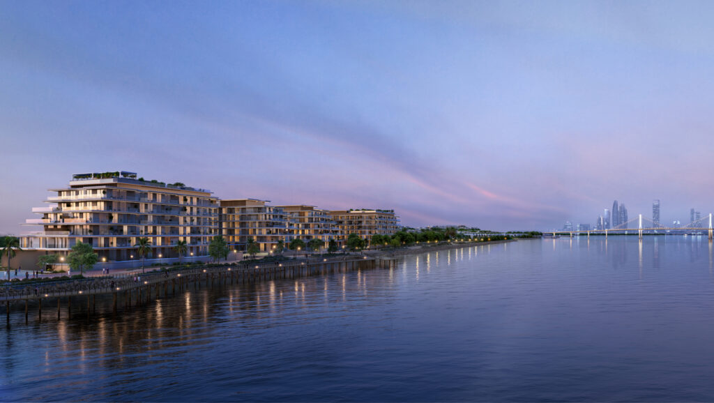 Une rangée d'immeubles modernes avec balcons borde une étendue d'eau calme au coucher du soleil, offrant un aperçu qui vous invite à tout savoir sur Modon Properties, avec une ligne d'horizon de la ville et un pont au loin sous un ciel pastel.