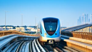 Une rame moderne du métro Gold Line de Dubaï, bleue et argentée, circule sur des voies surélevées, avec en arrière-plan la ligne d'horizon de la ville et des quartiers animés, sous un ciel bleu limpide.