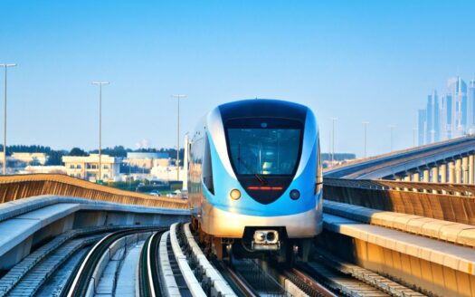 Une rame moderne du métro Gold Line de Dubaï, bleue et argentée, circule sur des voies surélevées, avec en arrière-plan la ligne d'horizon de la ville et des quartiers animés, sous un ciel bleu limpide.
