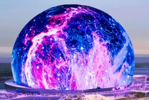 Un grand bâtiment sphérique comme la Sphère de Las Vegas est illuminé par des lumières tourbillonnantes bleues, roses et violettes. Des foules de personnes sont rassemblées autour de sa base et le ciel en arrière-plan apparaît au crépuscule.