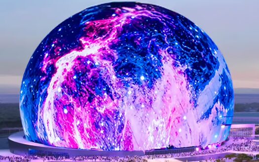 Un grand bâtiment sphérique comme la Sphère de Las Vegas est illuminé par des lumières tourbillonnantes bleues, roses et violettes. Des foules de personnes sont rassemblées autour de sa base et le ciel en arrière-plan apparaît au crépuscule.