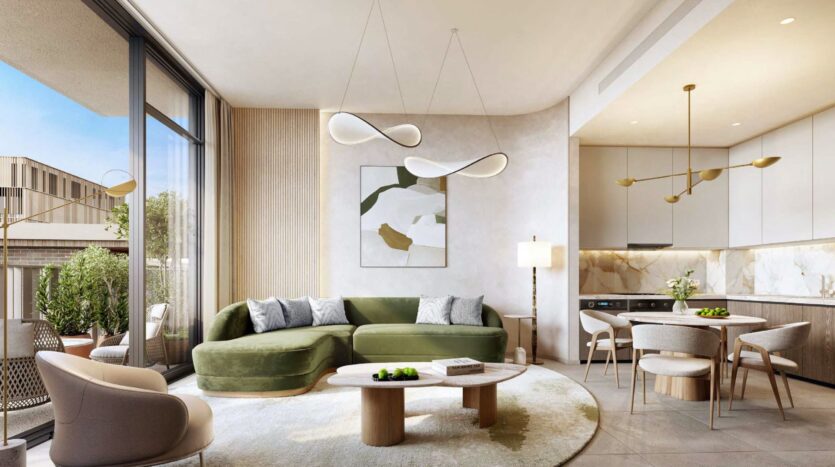 Le salon moderne et ensoleillé d'Arancia Yards by Beyond est composé d'un canapé sectionnel vert olive, d'une table basse ronde, d'œuvres d'art murales abstraites et d'une cuisine ouverte. Des fenêtres allant du sol au plafond révèlent un balcon avec des sièges extérieurs et de la verdure.