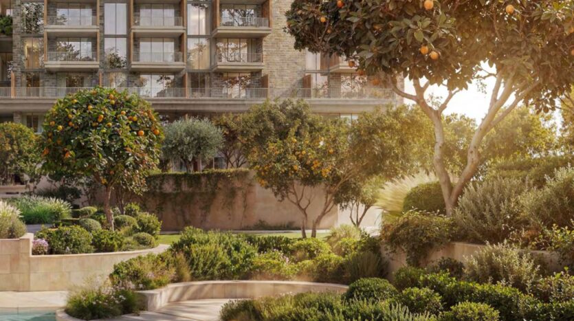 Un jardin paysager avec des buissons verdoyants et plusieurs orangers devant l'immeuble moderne Arancia Yards by Beyond, baigné par la lumière chaude du soleil.
