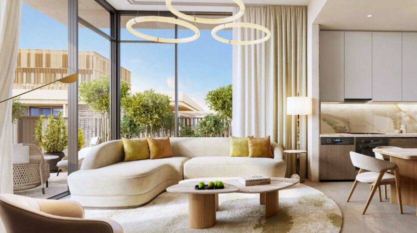 Un salon moderne à Arancia Yards by Beyond comprend un canapé beige incurvé, une table basse ronde en bois et de grandes fenêtres encadrant de la verdure. Des lampes suspendues élégantes ornent le plafond, mettant en valeur les tons neutres et la lumière naturelle dans tout l'espace.