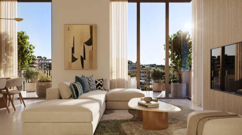 Le salon moderne de l'Arancia Yards by Beyond se compose d'un grand canapé sectionnel crème, d'œuvres d'art murales abstraites, de coussins décoratifs, d'une table basse en bois, d'une télévision et de la lumière du soleil qui traverse les grandes fenêtres pour atteindre un balcon avec des plantes à l'extérieur.