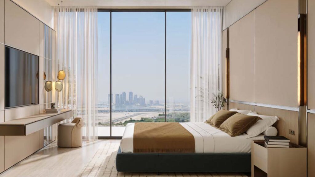 Chambre moderne à Binghatti Wraith, décorée dans des tons beiges et marron, avec un grand lit, une télévision, un bureau et une baie vitrée offrant une vue imprenable sur les toits de la ville et le front de mer. La lumière naturelle inonde cet espace minimaliste et élégant.