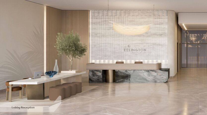 Le hall d'accueil moderne de l'Everly Place by Ellington se distingue par son sol en marbre, une longue table blanche accompagnée de tabourets marron, un arbre en pot et un comptoir d'accueil arborant le logo d'Ellington Properties. Un éclairage tamisé et des tons neutres viennent rehausser l'atmosphère accueillante.