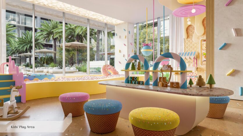 L'aire de jeux pour enfants d'Everly Place by Ellington, lumineuse et moderne, est équipée de mobilier aux couleurs pastel, de tabourets ronds, d'une table de jeux, d'un mur d'escalade, d'un cheval à bascule et de grandes baies vitrées donnant sur des palmiers. La décoration ludique et l'éclairage tamisé créent un espace joyeux.