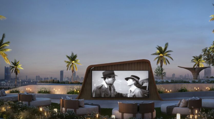 Un cinéma en plein air sur le toit, au coucher du soleil, aux Mövenpick Residences : un grand écran diffuse un film en noir et blanc, entouré de sièges confortables, de palmiers, avec la silhouette de la ville en arrière-plan. De petites lumières créent une ambiance chaleureuse et accueillante.