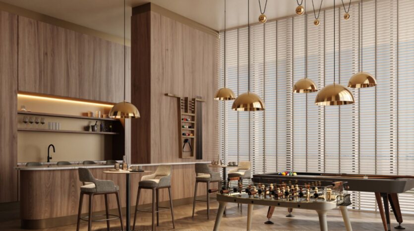 Salle de jeux moderne au Mövenpick Residences, avec une décoration en bois, un baby-foot, un îlot de cuisine et des tabourets de bar, des suspensions dorées, des étagères murales où sont exposés des verres, ainsi que de grandes fenêtres équipées de stores laissant entrer la lumière naturelle.