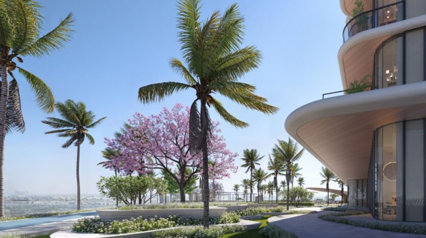 Un immeuble moderne à l'architecture courbe, situé au bord d'un jardin paysager agrémenté de palmiers, d'un arbre à fleurs roses et offrant une vue sur l'eau sous un ciel d'un bleu limpide : venez découvrir la sérénité de la vie aux Mövenpick Residences.