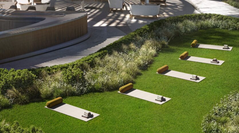 Cinq tapis de yoga, accompagnés de coussins jaunes et de serviettes pliées, sont soigneusement disposés sur une pelouse impeccablement entretenue aux Mövenpick Residences, à côté d'arbustes verdoyants, avec des sièges d'extérieur modernes et une structure incurvée en arrière-plan.