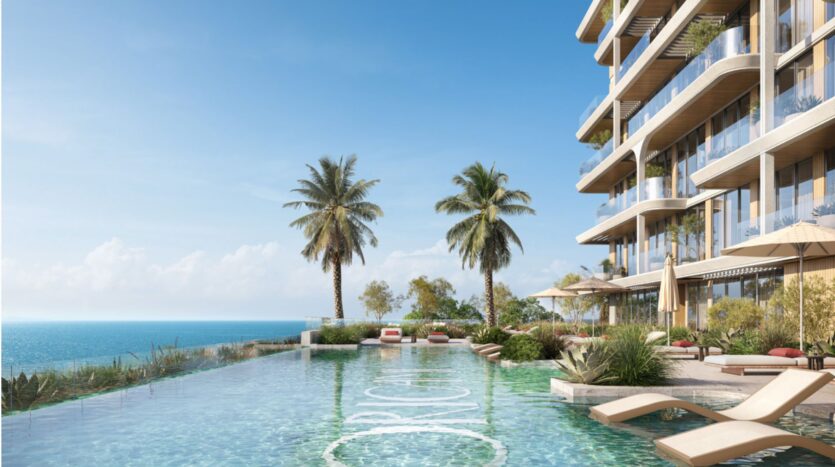 Une piscine à débordement donnant sur l'océan, bordée de palmiers et de transats, avec, sur la droite, sous un ciel bleu limpide, les Soma Residences, un immeuble moderne de plusieurs étages doté de balcons.