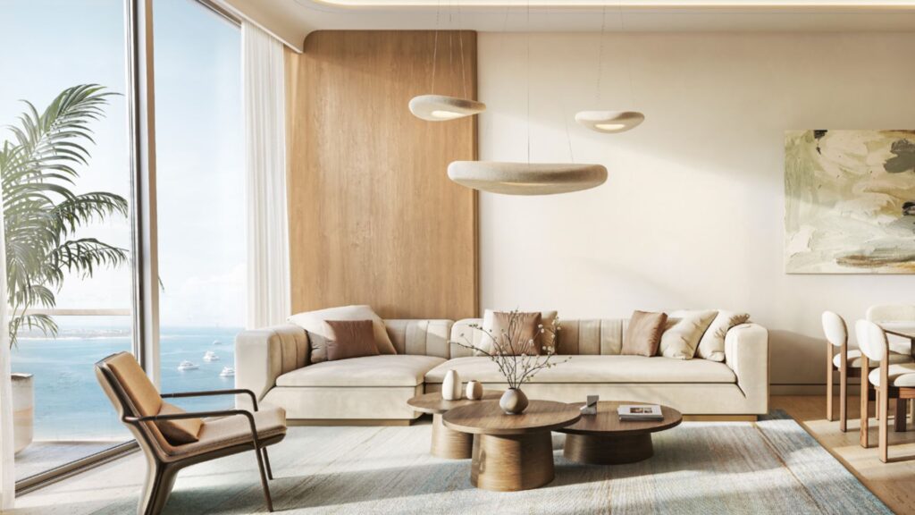 Le salon moderne des Soma Residences est aménagé avec un canapé beige, des tables basses en bois et un fauteuil. De grandes baies vitrées offrent une vue sur l'océan. Des tons neutres, des œuvres d'art abstraites accrochées aux murs et des suspensions sculpturales viennent parachever l'espace.