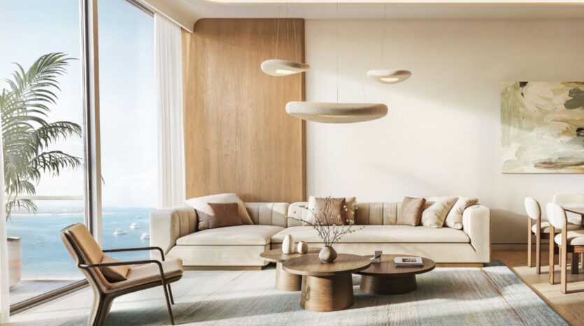Le salon moderne des Soma Residences est aménagé avec un canapé beige, des tables basses en bois et un fauteuil. De grandes baies vitrées offrent une vue sur l'océan. Des tons neutres, des œuvres d'art abstraites accrochées aux murs et des suspensions sculpturales viennent parachever l'espace.