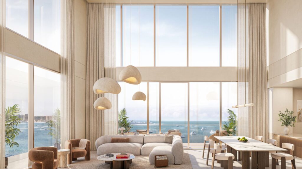 Salon spacieux et moderne au sein des Soma Residences, avec de hauts plafonds, de grandes baies vitrées donnant sur l'océan, un mobilier beige, des suspensions, des plantes vertes et une table à manger en bois, le tout dans un cadre lumineux et aéré.